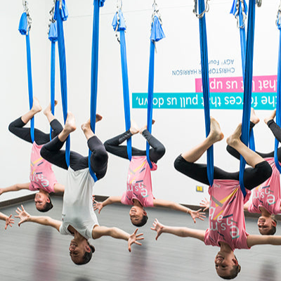 Group of people in blue Antigravity Fitness aerial Yoga hammocks in a room with a white wall and visible branding.