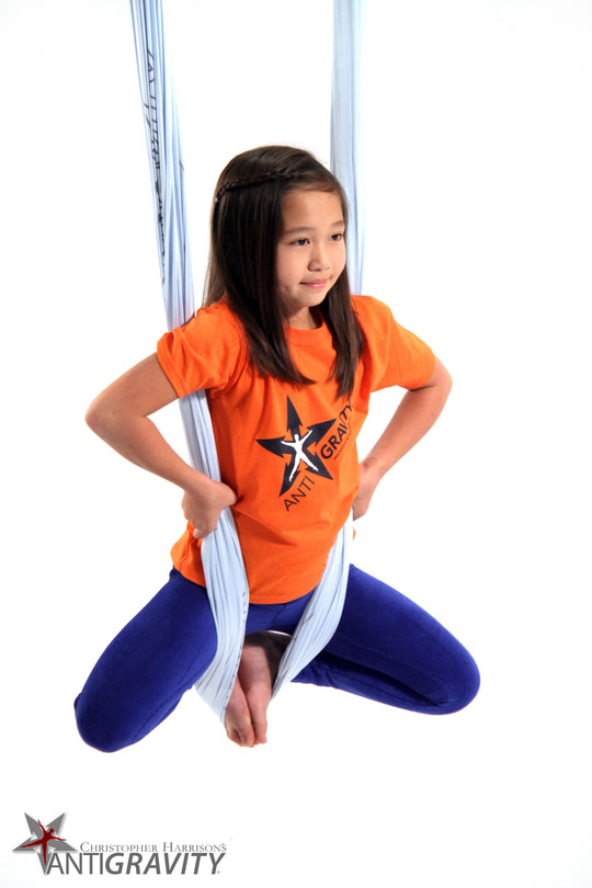 Child in orange shirt and blue pants sitting on a white aerial silks with Antigravity logo.